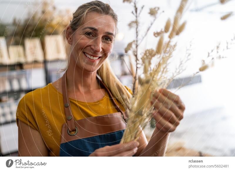 A smiling female florist holds a bunch of dried wheat in a bright workshop cheerful floristry blonde hair apron yellow shirt craftsmanship floral design