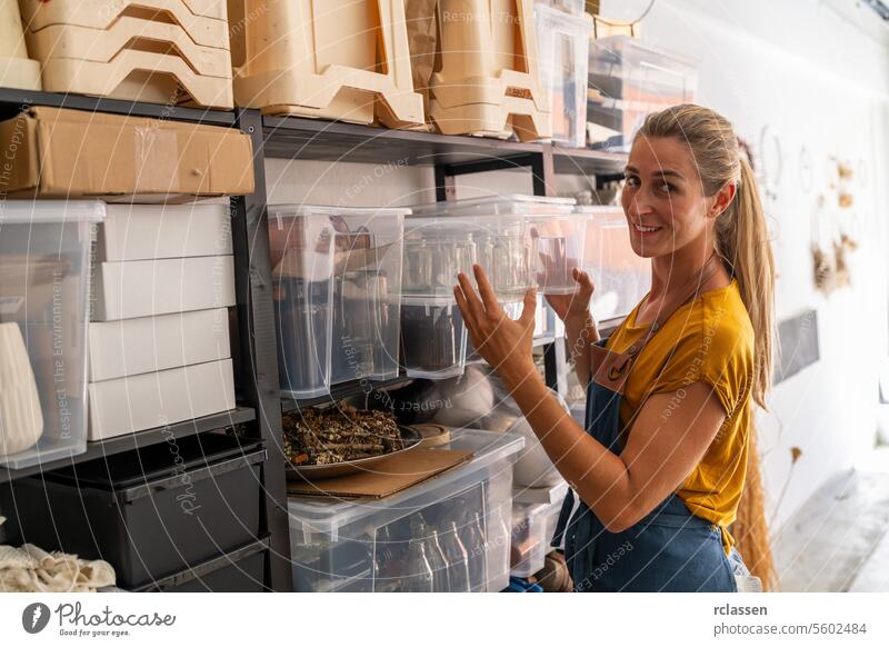 woman in a mustard-colored shirt and denim apron is arranging clear storage containers on shelves in a dried flowers workshop, smiling as she looks at the camera