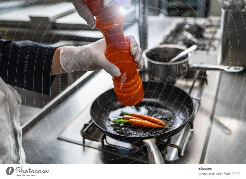 Chef in hotel or restaurant kitchen cooking and seasoning fried carrots in the frying pan with a pepper or salt mill. Luxury cooking concept image man