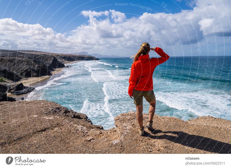 Hiker in red jacket looking out at the ocean waves from a cliff on a sunny day fuerteventura hiker seascape travel adventure nature coastline exploration