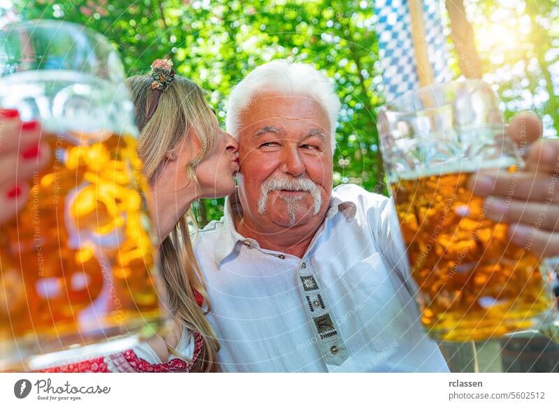 daughter gives her father a kiss on the cheek and say cheers or clinking glasses with mug of beer in Bavarian beer garden or oktoberfest pensioner bavarian flag