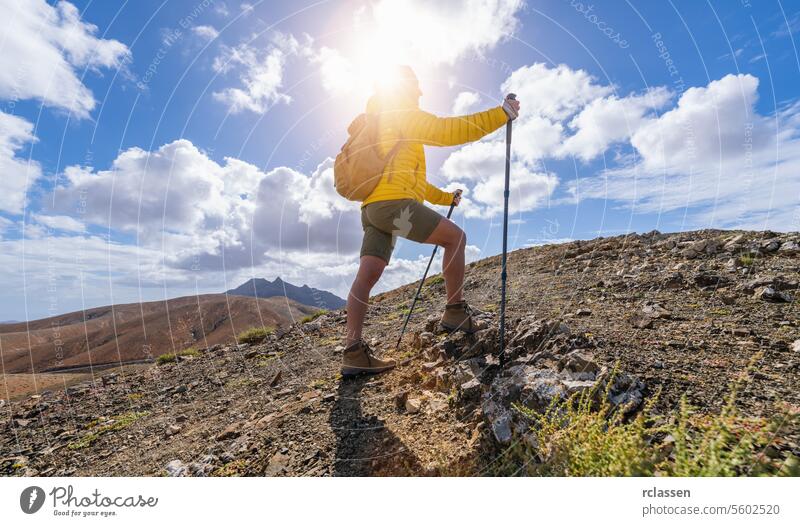 Hiker in yellow jacket climbing a mountain path against a sunny, cloud-filled sky fuerteventura hiker sunny sky clouds hiking trekking pole outdoor adventure