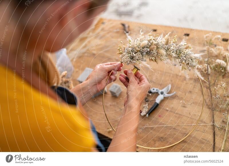 Close-up of a florist's hands wiring dried flowers onto a hoop against a wooden table backdrop close-up crafting floral design flower arrangement workshop