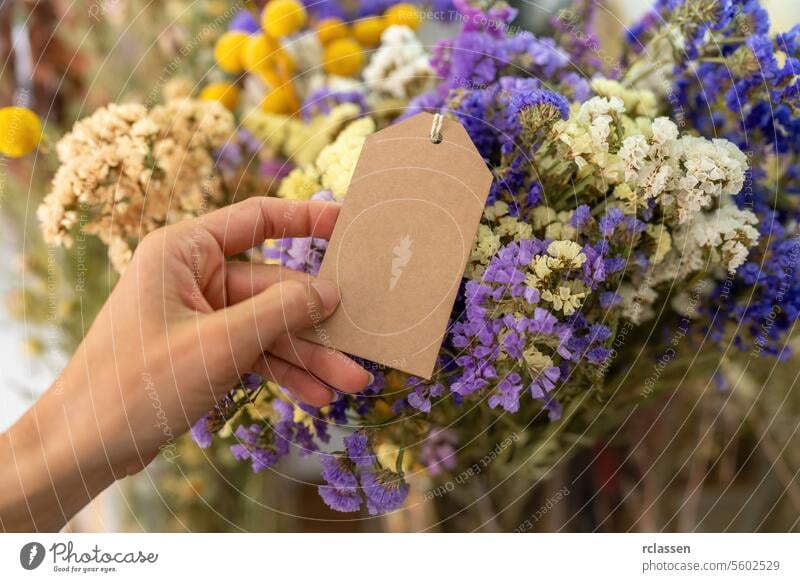 Hand holding a blank brown tag in front of a bouquet of colorful dried flowers, with copyspace for your individual text. decorative autumn florist floral