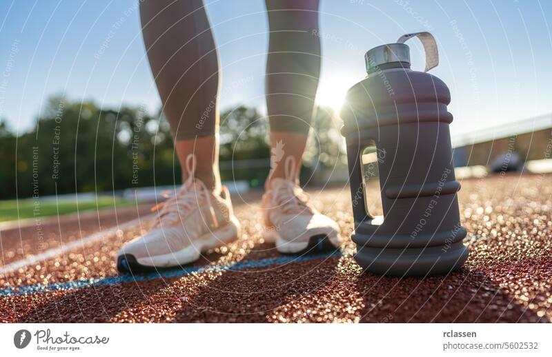 Close-up of a large water bottle on a running track with woman's legs in background close-up fitness hydration sportswear athlete training workout health