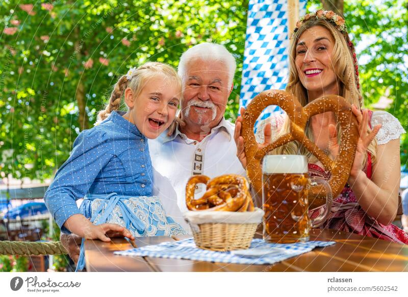 Happy family in proper Tracht holding big pretzel and drinking in Bavarian beer garden at Oktoberfest, folk or beer festival in germany girl kid woman