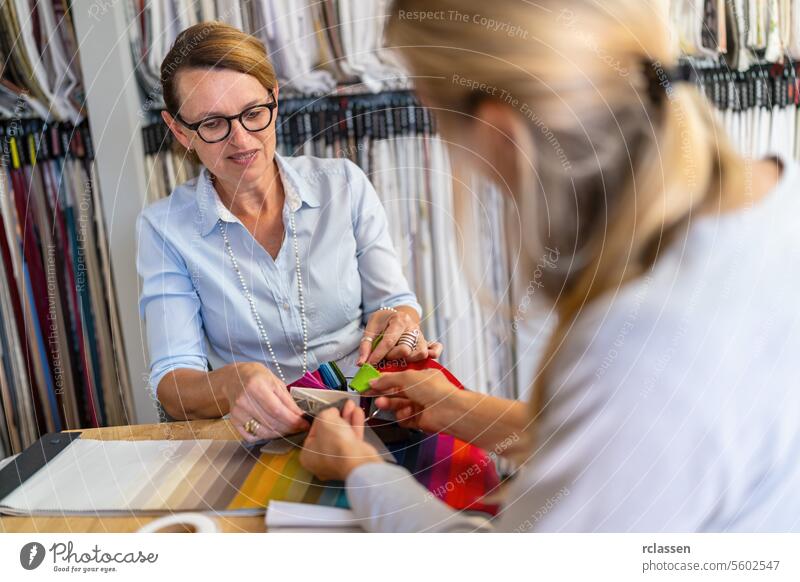 Seamstress sits with her customer in the shop and advises her on choosing her curtain fabric. Seamstress  smiling and touching a color swatch while the other, partly visible, listens attentively.
