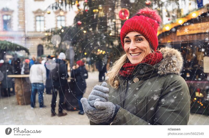 Smiling woman in red beanie and scarf with snowflakes at a festive Christmas market at winter time in germany, with copyspace for your individual text.
