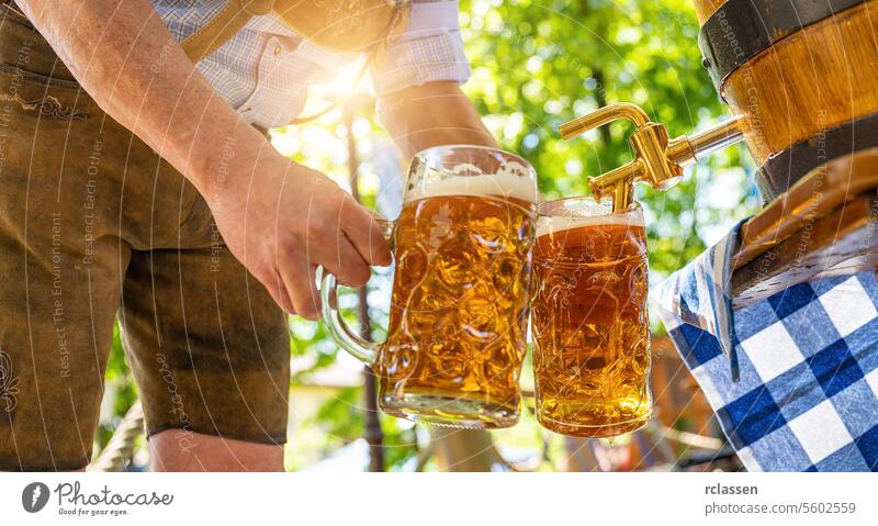 Bavarian man in leather trousers is pouring large lager beers in tap from wooden beer barrel in the beer garden. Background for Oktoberfest or Wiesn, folk or beer festival (German for: O’zapft is!)