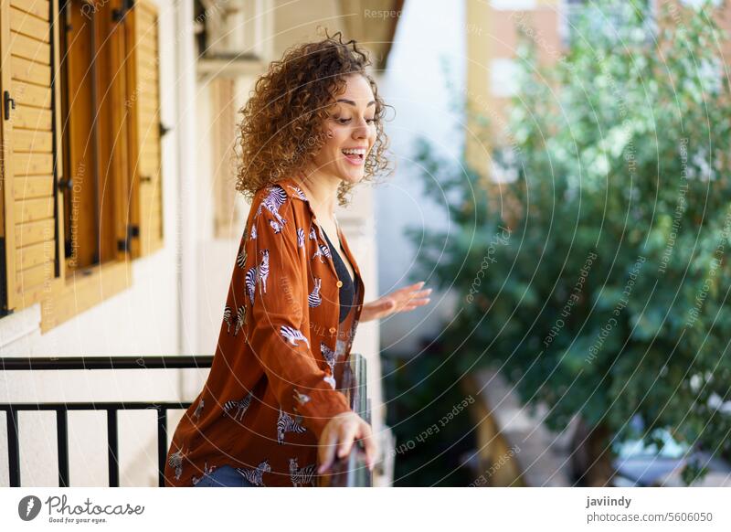 Joyful young brunette curly hair woman standing on balcony of modern building smile cheerful town happy content glad casual positive house pleasant alone