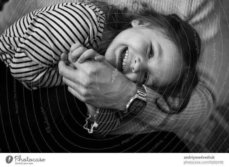 Happy children's day. Black and white photo of unrecognisable dad with adorable laughing little daughter are having fun spending time together on weekend at home. Playful child girl and daddy hugging