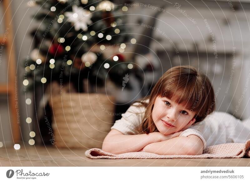 Little girl near decorated fir tree with lighting garlands in traditional holiday interior. Child Girl in warm clothes lying on floor and looking in camera. Winter family holiday concept. New year eve