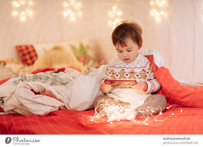 adorable little girls in worm clothes with winter ornaments are having fun, communicate and share secrets on a bed under blanket in a cozy bedroom decorated garlands, New Year's family holiday