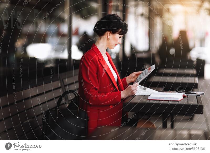 A attractive businesswoman in corporate outfit red blazer holding morning newspaper while sitting on cafe terrace. Manager is reading the latest world news during a coffee break at work