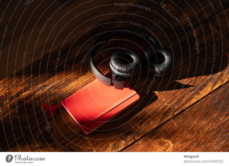 Black headphones lie in an atmospheric mood on a red notebook caught in the sunlight on dark brown grained wooden floorboards, ready to listen to the music and write down your thoughts