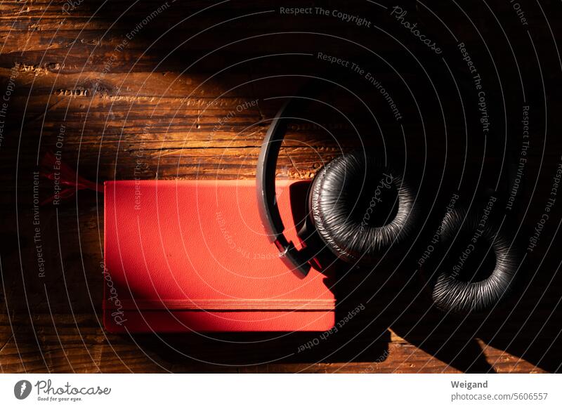 Black headphones lie in an atmospheric mood on a red notebook caught in the sunlight on a dark-grained wooden background, ready to listen to the music and write down your thoughts
