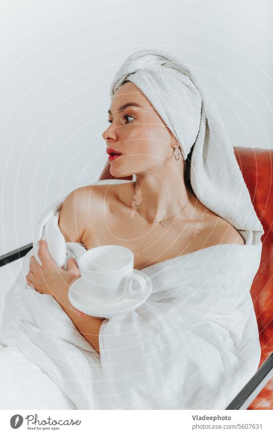 Woman drinking coffee or tea wrapped in a white blanket and towel around head. White background woman morning home lifestyle bathrobe bedding girl lady cozy