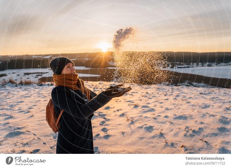 A woman in a snow-covered field tosses snow with her hands against the backdrop of a sunset sunset. winter warm clothes female people person European White