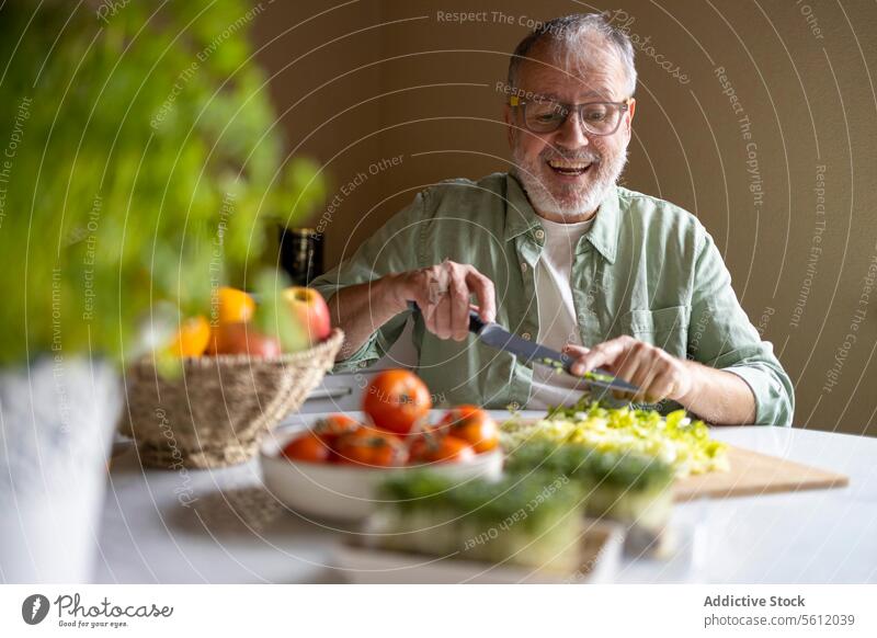 Cheerful senior man in glasses eyewear cutting lettuce in pieces with knife on wooden chopping board while preparing salad at home table kitchen smile