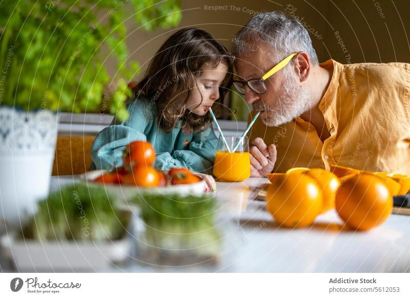 Senior man and girl drinking fruit juice on table grandfather granddaughter orange straw glass sharing senior cute family home selective focus people lifestyle