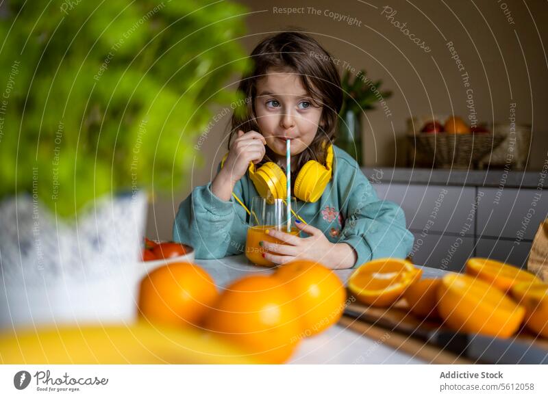 Smiling adorable girl with headphones on neck and looking away while drinking orange juice on table at home smiling glass straw selective focus lifestyle