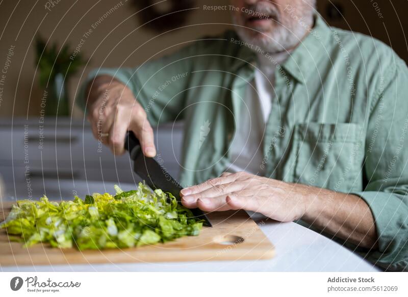 Crop hands of anonymous senior man cutting lettuce in pieces with knife on wooden chopping board while preparing salad at home crop table kitchen caucasian