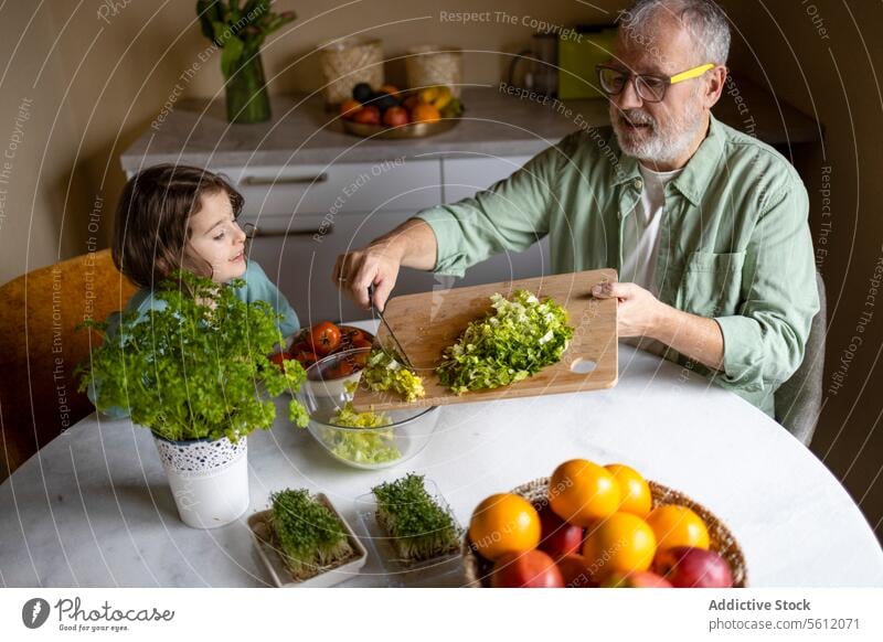 Man preparing vegetable salad with kid at table grandfather granddaughter girl senior man fruit plant herb chopping board bowl knife learning elderly lettuce