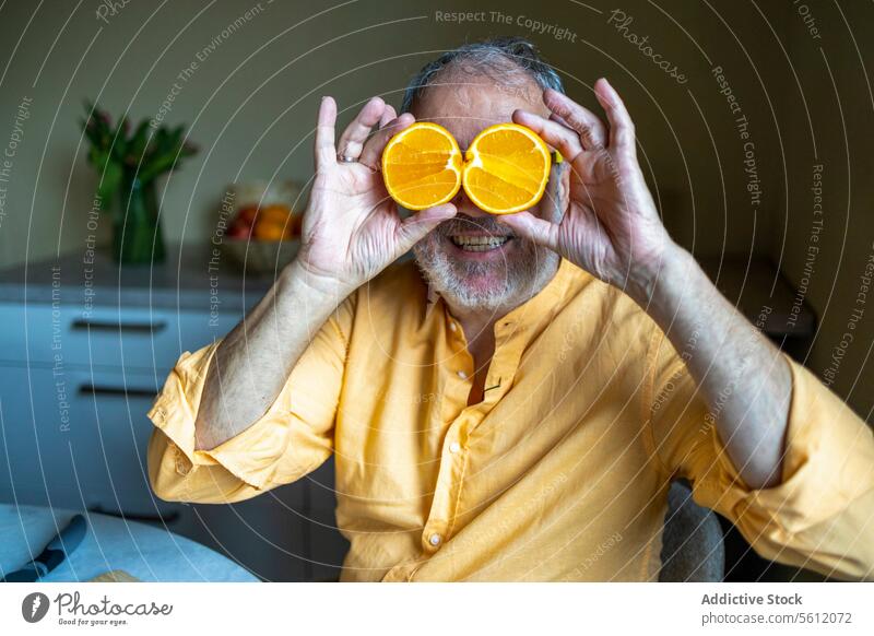 Aged man covering eyes with orange slices at home senior smiling halved cheerful kitchen sitting casual attire lifestyle aged fruit caucasian blurred background