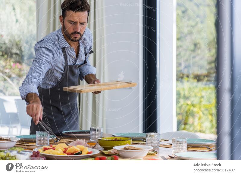 Bearded Male cook in apron serving appetizers on table in modern living room man chef serve breakfast dining room workday homemade appetizing male tongs