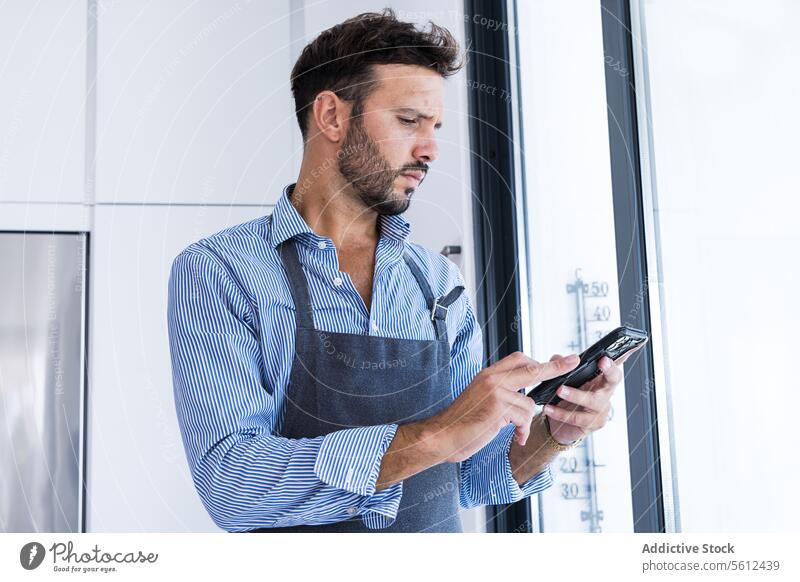 Side view of focused looking man in apron browsing smartphone in daylight near window chef using communicate online chat text message room male adult beard