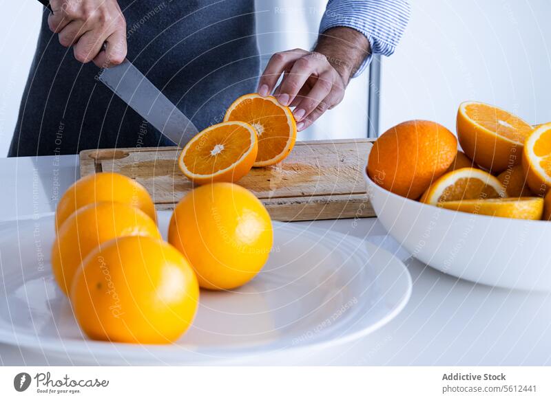 Unrecognizable crop man chef in apron standing and cutting oranges on chopping board in modern kitchen knife half fruit organic fresh male sleeve slice prepare