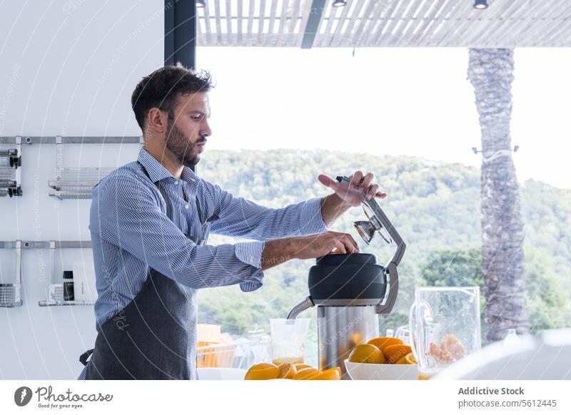 Side view of bearded man in apron standing near table with orange fruits squeezer in modern kitchen while preparing orange juice chef prepare breakfast cut male