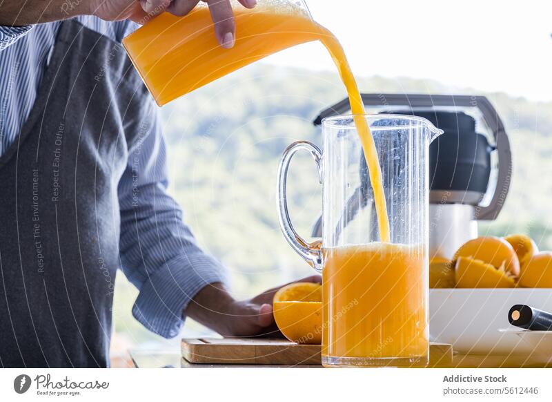 Unrecognizable chef in apron standing at kitchen table and pouring orange juice into jug in modern kitchen man breakfast squeeze fresh male sleeve prepare drink