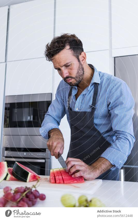 Private bearded chef in apron standing at table while slicing watermelon in kitchen man cook cut slice knife healthy food vitamin breakfast male modern