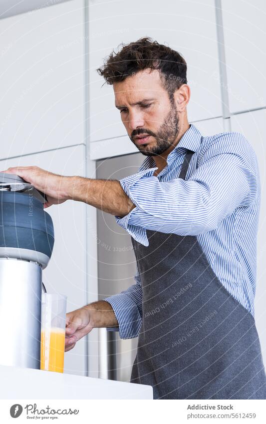 Low angle of adult man in apron standing near table with orange fruits squeezer in modern kitchen chef prepare breakfast juice male beard sleeve organic fresh