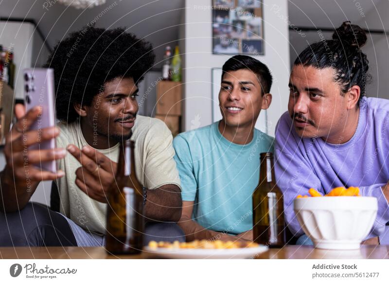 Male friends see a photo in smartphone at home smile afro hair leisure together living room friendship weekend mobile beer bottle looking cellphone social media