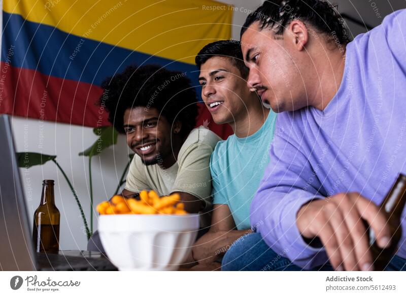 Happy male fans watching match over computer at home friends smile laptop game sport excited beer bottle snack sit colombian flag drink beverage food happy bowl