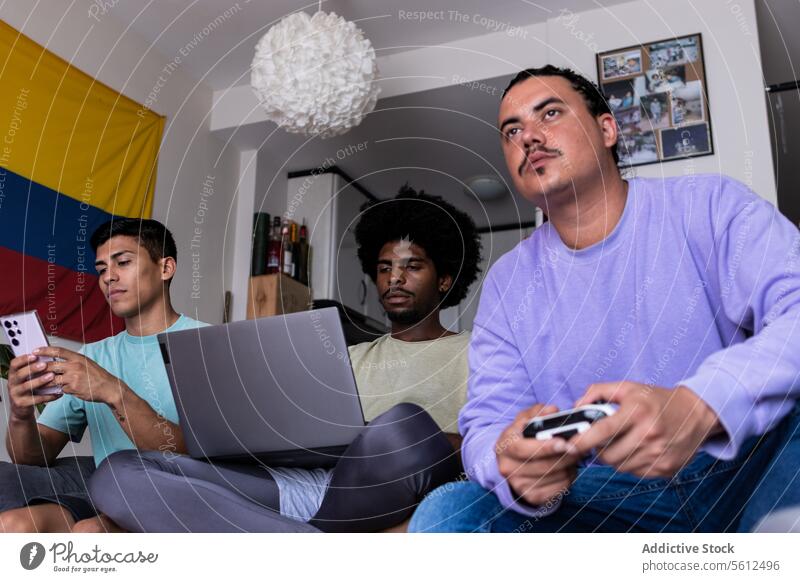 Multiracial male friends using technology at home smartphone laptop console pointing looking at screen computer table living room weekend diverse messaging