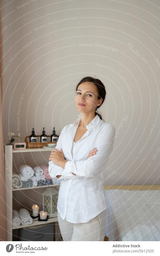 Serene massage therapist in spa center masseuse arms crossed confident beautiful portrait positive uniform white smile looking at camera stand bed salon woman