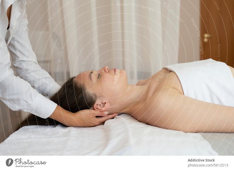 Side view of female customer wrapped in white towel with closed eyes lying on bed and receiving neck therapy massage from unrecognizable message therapist