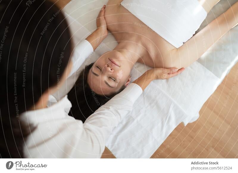 Top view of female customer wrapped in white towel with closed eyes lying on bed and receiving neck therapy massage from unrecognizable message therapist client