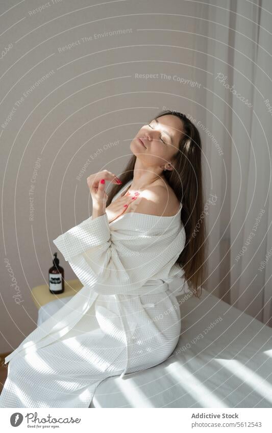Positive calm mid adult woman in white bathrobe with eyes closed touching body and relaxing while sitting on bed after spa procedures positive rest content