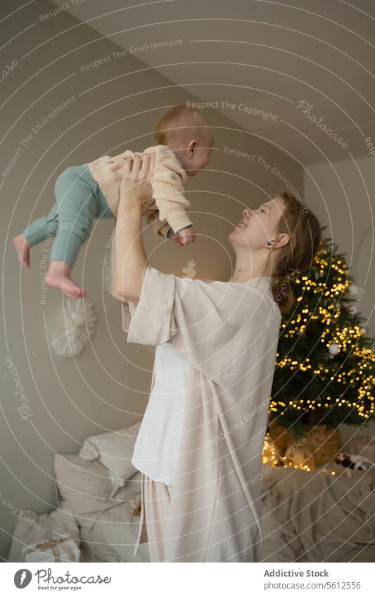 Young mother lifting baby with Christmas tree backdrop christmas home joy tender warm light cozy background indoors celebration festive woman child infant