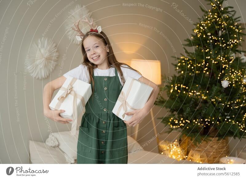 Happy girl holding gifts with Christmas tree backdrop christmas present holiday decoration lights smiling reindeer antlers happy festive season joy celebration