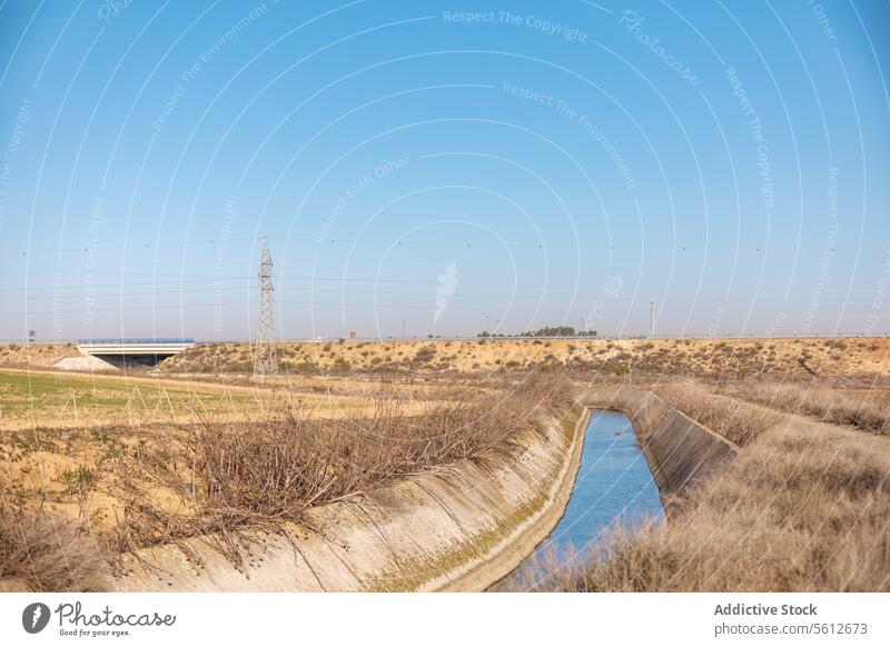 Canal in arid landscape with electricity pylon and highway dry barren canal irrigation water energy infrastructure transmission tower sky blue nature