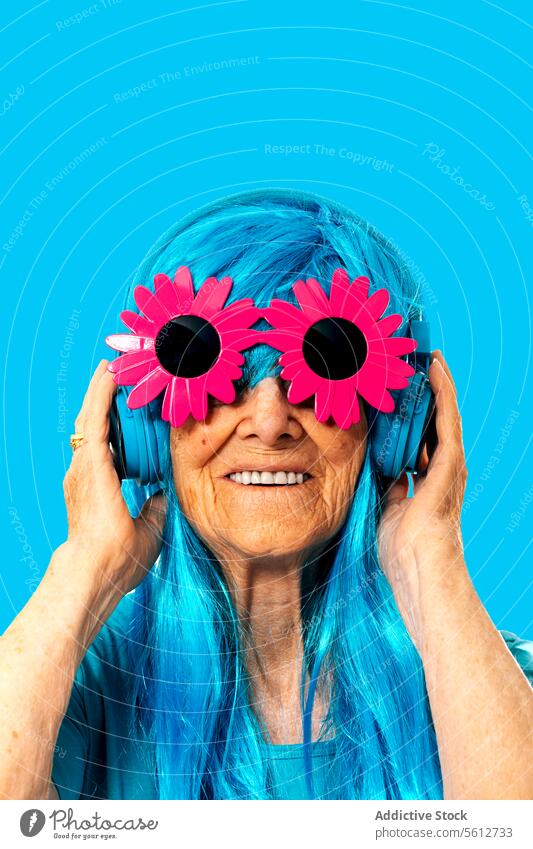 Portrait of happy elderly female in blue wig with headphones and wearing flower frame sunglasses against blue background woman portrait wrinkle appearance smile