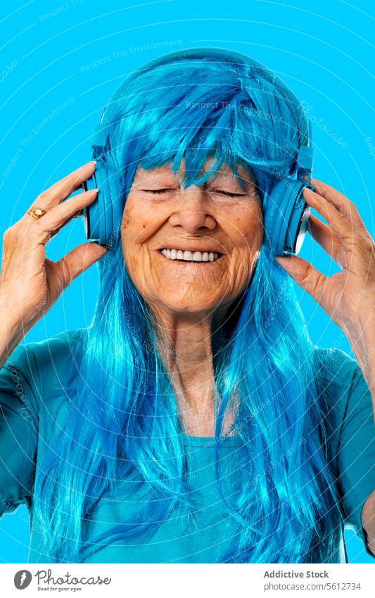 Cheerful aged woman listening to music via headphones with closed eyes on blue background portrait enjoy song wig color match smile happy cheerful studio shot