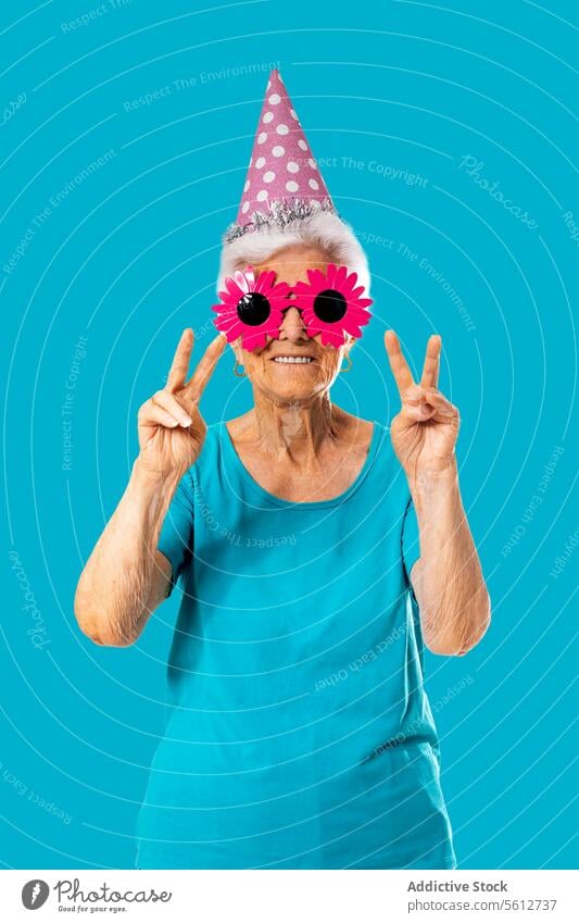 Funny aged woman with funny pink sunglasses and birthday cap showing two fingers in studio gesture peace celebrate smile happy cheerful female senior elderly