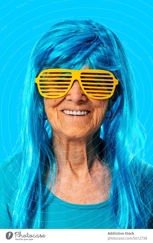 Smiling elderly woman in sunglasses and blue wig looking at camera while standing on blue background portrait stripe retire pensioner funny wrinkle accessory
