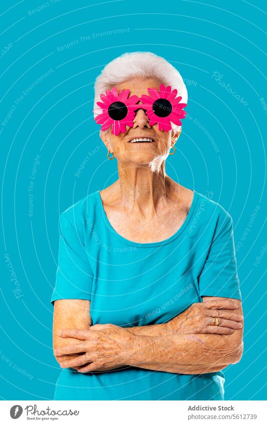 Cheerful elderly woman wearing funny pink sunglasses while standing with crossed arms on blue background arms crossed childish retire pensioner accessory